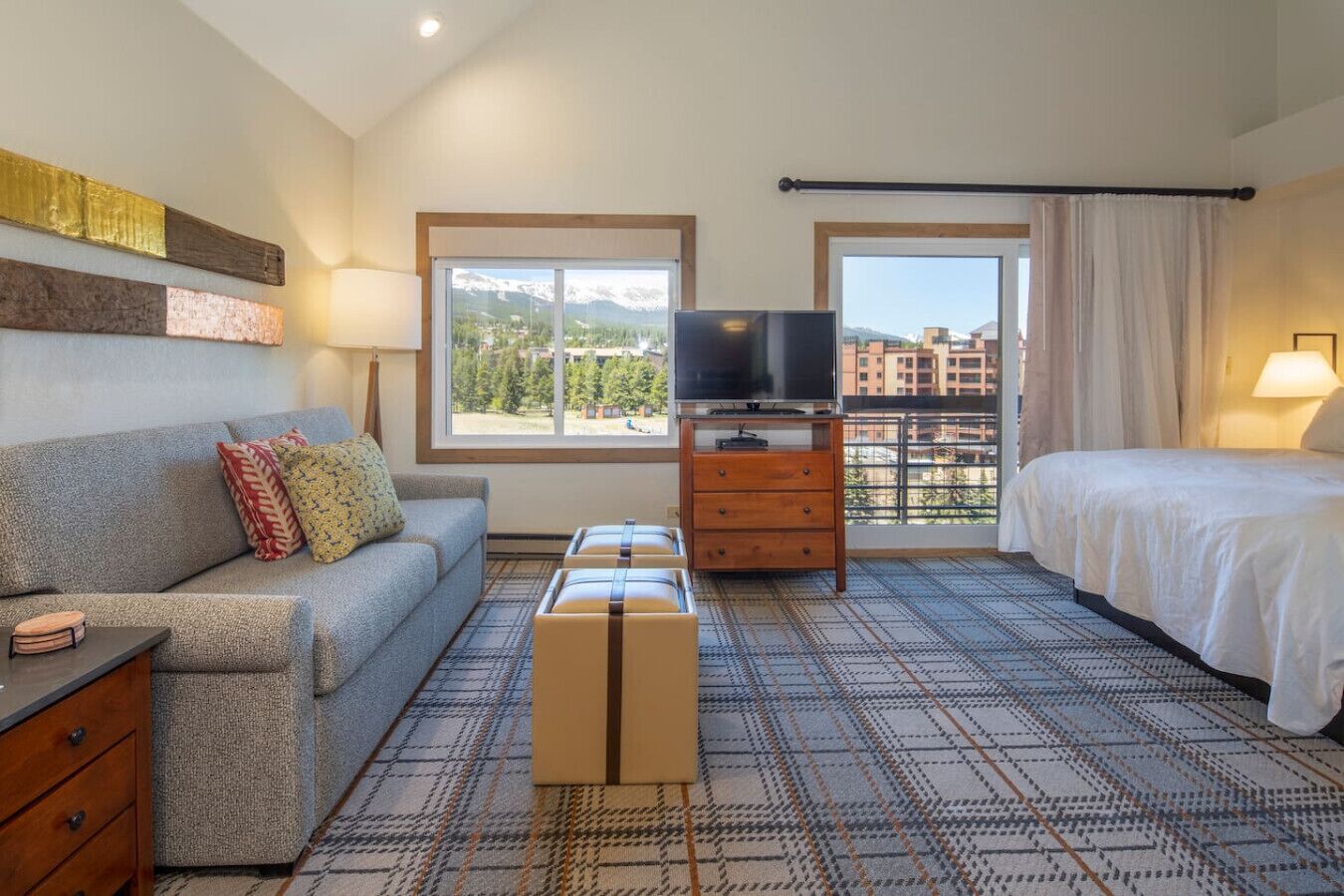 Ein Hotelzimmer mit Sofa, Bett, Fernseher, Kommode und einem Fenster mit Blick auf Gebäude und Berge.