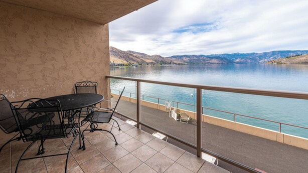 Un balcon avec une table et des chaises donnant sur un lac, avec des montagnes en arrière-plan.