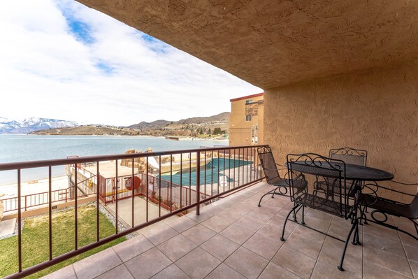 A balcony with a view of a beach and mountains, a pool, and outdoor furniture.