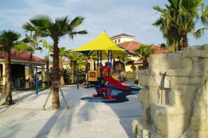 Children can play on the outdoor play structure with a slide surrounded by palm trees and a rock formation.