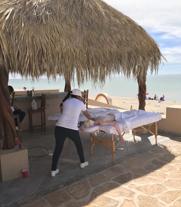 Ett område för massagebehandlingar med ett massageställ av trä, ett halmtak och utsikt över stranden.