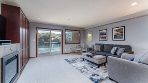 A spacious living room with a gray sofa, a coffee table, and a fireplace. There is a large window with a view of hills and a balcony.