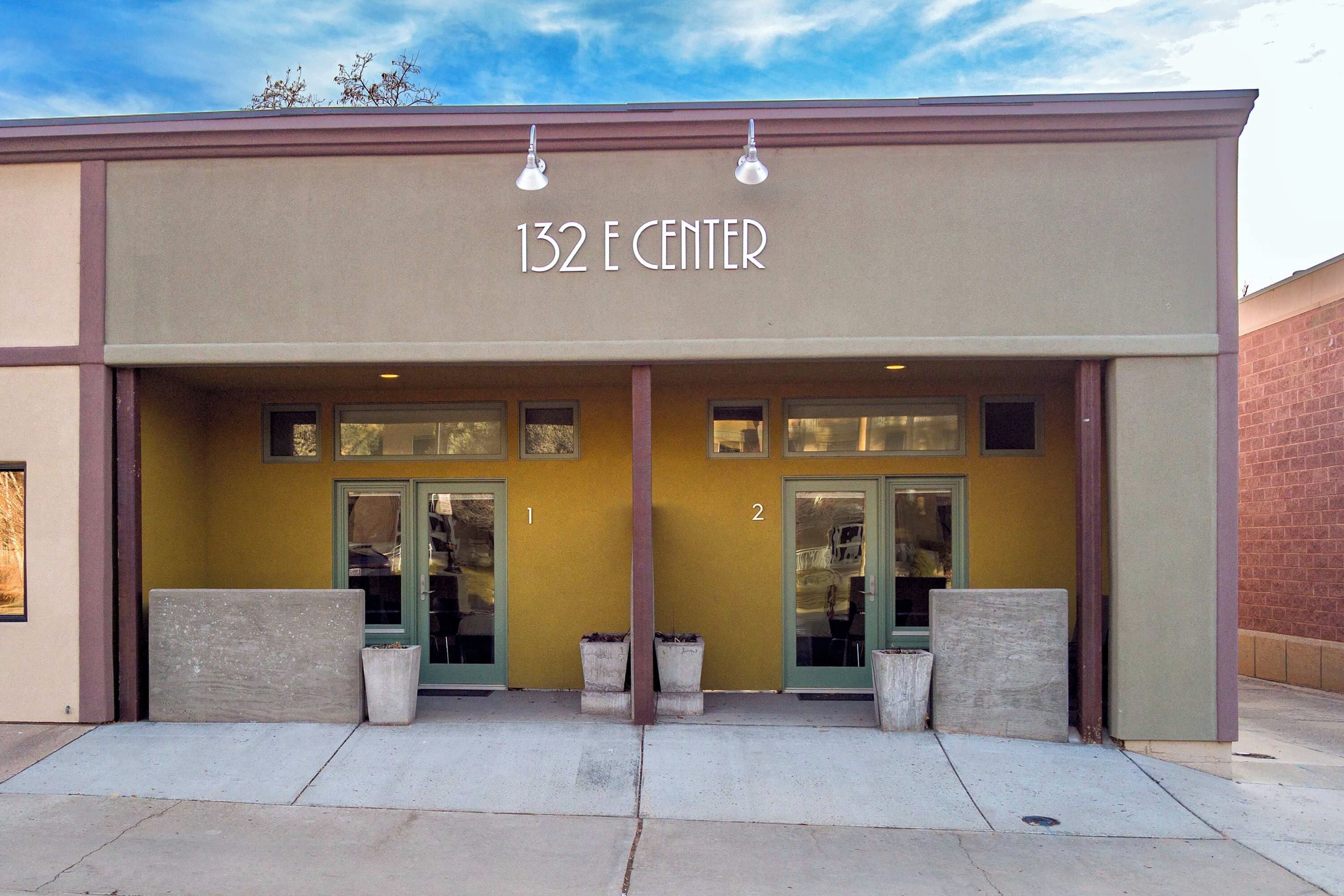 Ein Gebäude mit dem Schild "132 E CENTER" über dem Eingang. Es gibt zwei Türen, jeweils mit einem Fenster darüber. Die Fassade ist beige mit braunen Verzierungen.