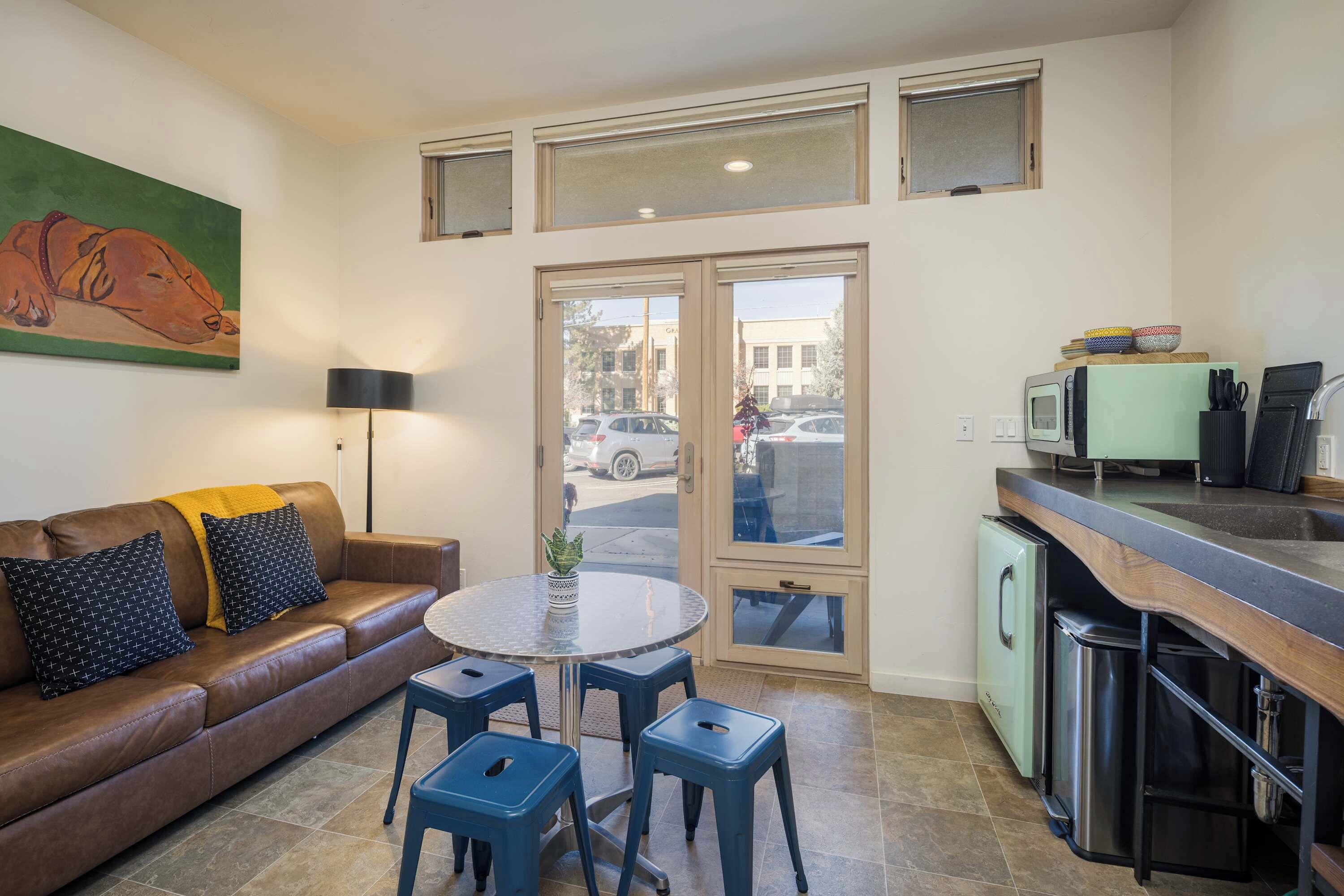 Un espacio habitable compacto con sofá, una mesa redonda y taburetes azules. Se observa una zona de cocina con refrigerador, microondas y lavamanos. Un cuadro de un perro adorna la pared.