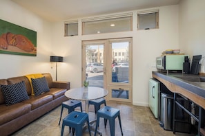 A compact living space with a sofa, a round table, and blue stools. A kitchen area is visible with a refrigerator, a microwave, and a sink. A painting of a dog decorates the wall.
