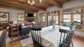 A rustic interior with a dining table, chairs, a fireplace, and large windows.