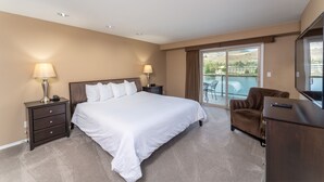 A bedroom with a large bed, two bedside tables with lamps, a brown armchair, a dresser, and a balcony with a view of the mountains.