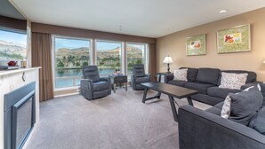 A modern living room with a sofa, chairs, a coffee table, and a fireplace. Large windows offer a view of a lake and mountains.