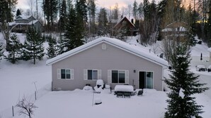 Uma casa térrea com telhado e quintal cobertos de neve, cercada por altas árvores perenes.