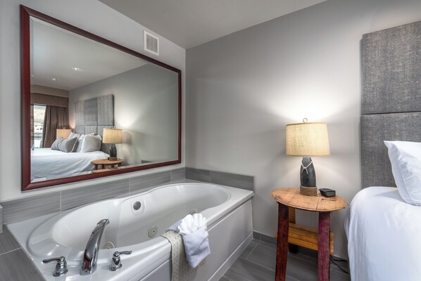 A modern hotel room with a large bathtub, a wooden side table with a lamp, and a large mirror reflecting the room.