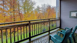 A wooden deck with a railing, green plastic chairs, and a view of autumn trees. - Spacious ski-in, ski-out 2-BR Jay Peak Village Condo, right on trail. A/C incl.! (Jay)