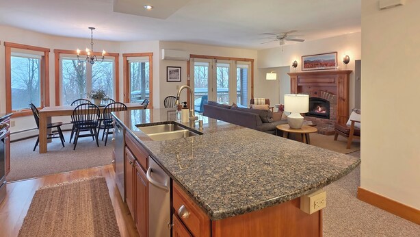 A spacious kitchen with granite countertops, wooden cabinets, and a central island. There is a dining area with a table and chairs, and a fireplace with a mantel. - Spacious ski-in, ski-out 2-BR Jay Peak Village Condo, right on trail. A/C incl.! (Jay)