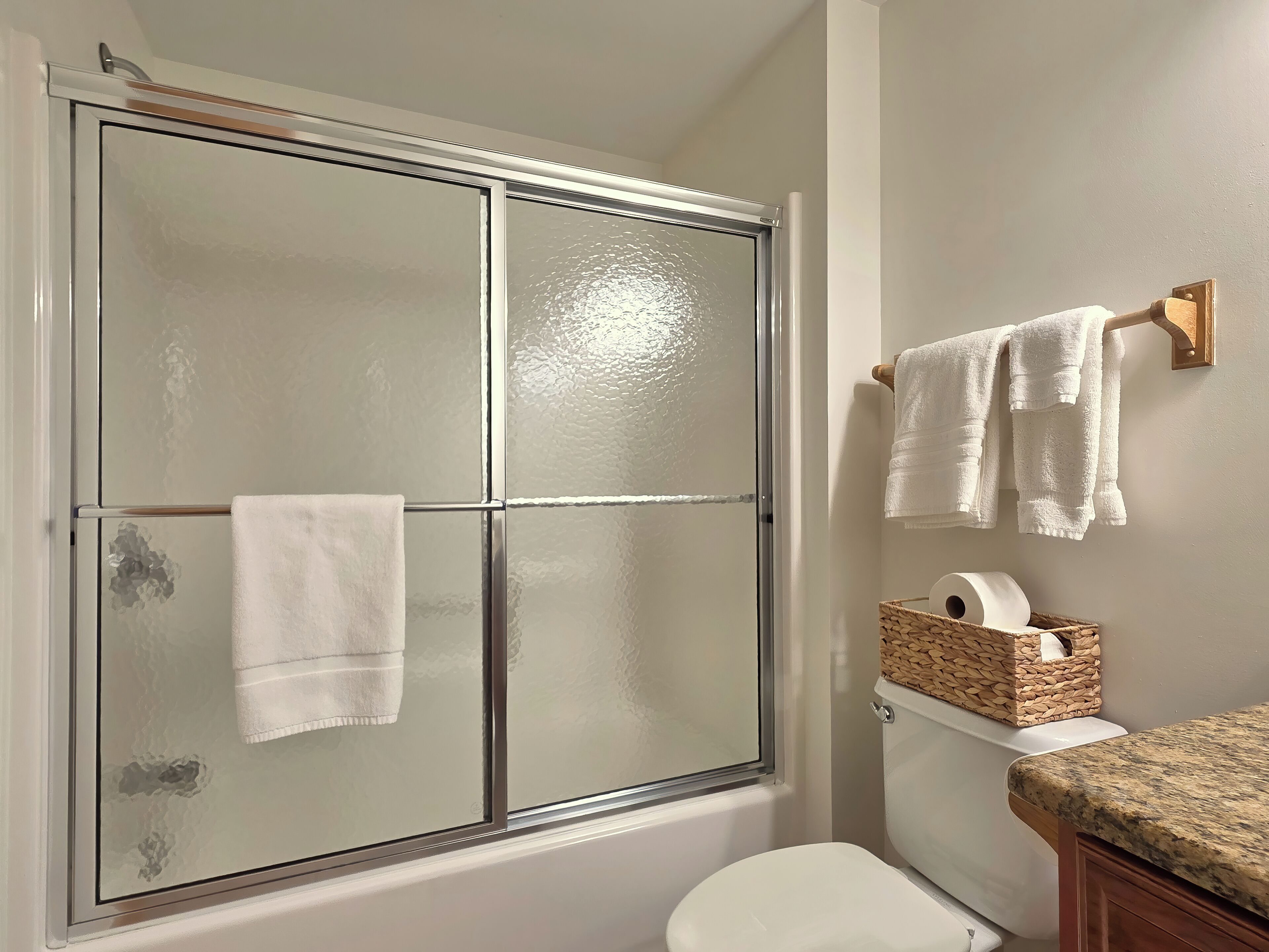 Un baño con cabina de ducha de vidrio, inodoro, lavamanos con encimera de granito y una cesta con un rollo de papel higiénico.