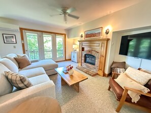 A living room with a fireplace, a sofa, a television, and a ceiling fan. - Spacious, Updated Ski-in, Ski-out 2-BR Jay Peak Village Condo, right on trail! (Jay)