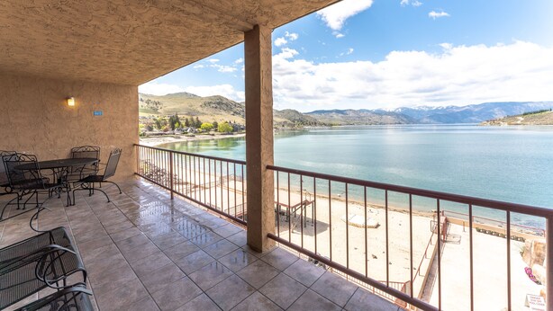 A covered outdoor area with a table and chairs, a view of a lake, and a mountainous landscape in the background. - GrandView 401 - 2 Bedroom Waterfront condo (Chelan)
