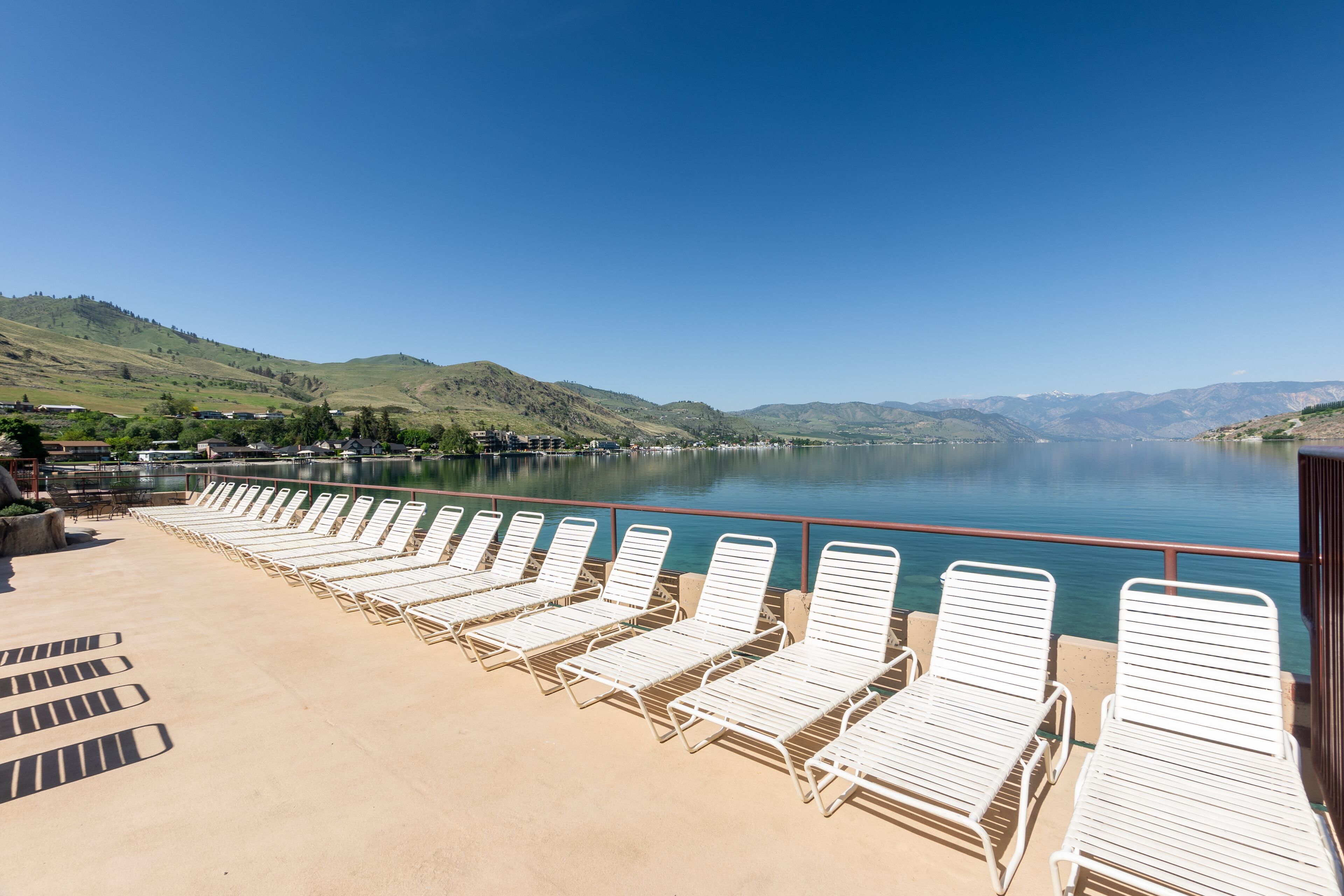 Une terrasse avec des chaises longues blanches donnant sur un lac paisible, le tout entouré de collines.