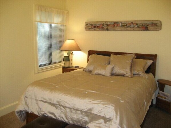 A neatly made bed with a beige comforter and pillows, a bedside lamp, a window with blinds, and a painting on the wall.