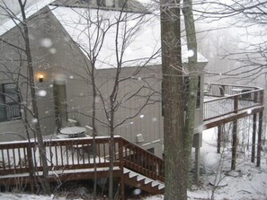 A snow-covered deck with a staircase leading to a second level, surrounded by bare trees and a building with a light fixture.