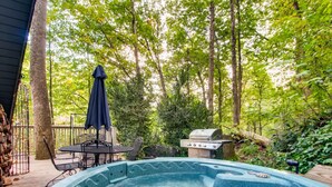 Ein Whirlpool, umgeben von einer Terrasse mit Gartenmöbeln, einem Grill und einer bewaldeten Kulisse.