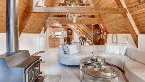 Ein gemütlicher Loftsraum mit Holzbalkendecke, einem modernen Glastisch, einer Couch und einem Kaminofen.
