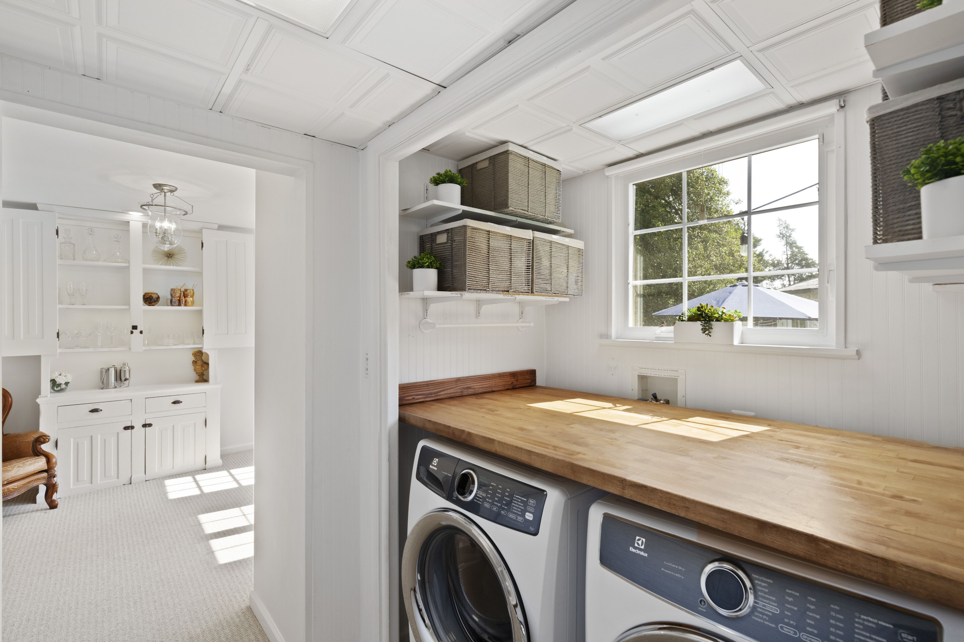 A laundry area with a washing machine, a dryer, and a wooden countertop. There are shelves with baskets and potted plants.