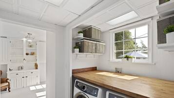 A laundry area with a washing machine, a dryer, and a wooden countertop. There are shelves with baskets and potted plants.