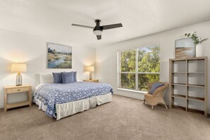 A bedroom with a bed, two bedside tables, a chair, a window with blinds, a ceiling fan, and artwork on the walls.