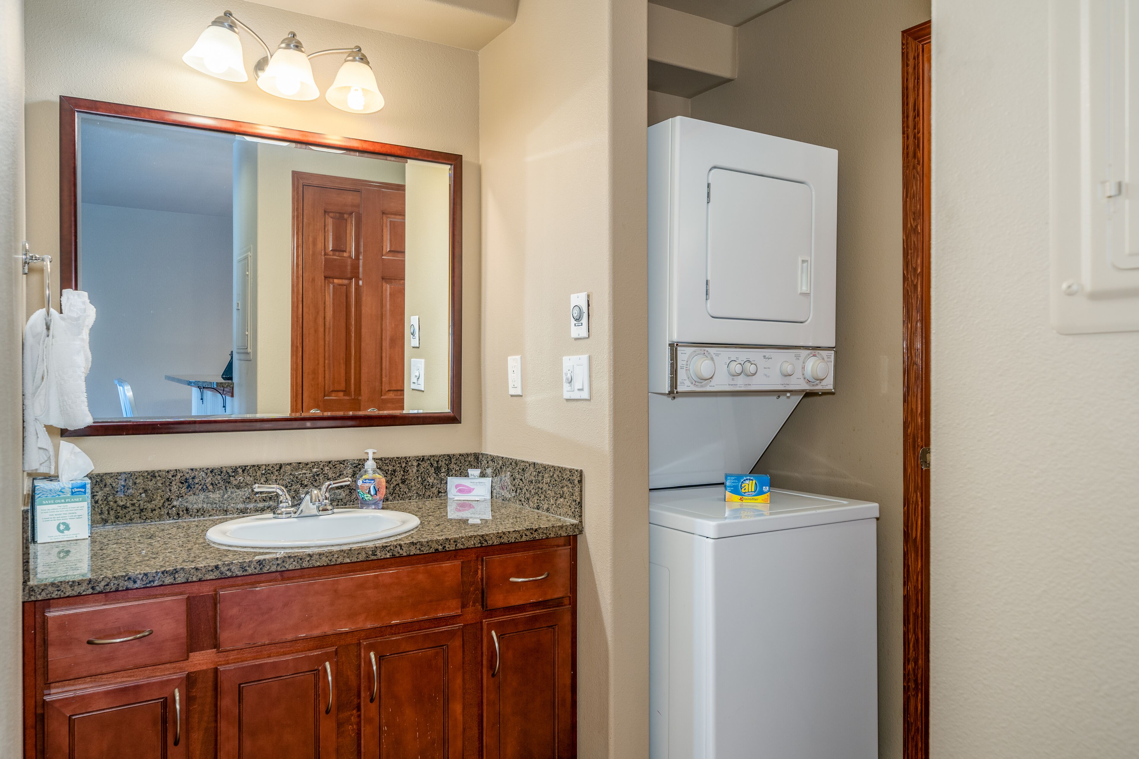 Une salle de bain avec un lavabo surmonté d’un plan de travail en granit, des meubles en bois, un miroir avec éclairage intégré, un lave-linge blanc et un porte-serviettes.