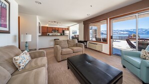 Amplia sala de estar con sofá, sillón y una mesa de centro. Se observa una cocina al fondo con armarios de madera y un microondas. La habitación tiene ventanas amplias que ofrecen vistas a un balcón con mobiliario exterior.