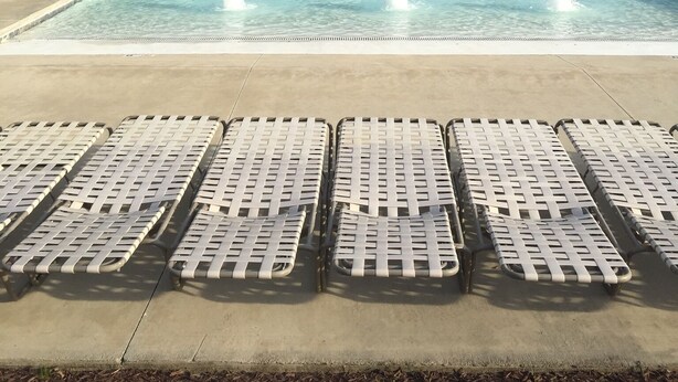 A rectangular swimming pool with clear blue water, surrounded by a concrete deck. There are lounge chairs aligned in the foreground.