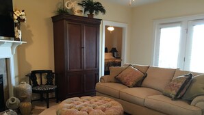 A living room with a beige sofa, a patterned ottoman, a dark wooden cabinet, and a chair with a patterned cushion.