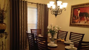 A dining area with a table set for four, surrounded by wooden chairs. A vase with white flowers is on the table, and a framed picture of a dog is on the wall.