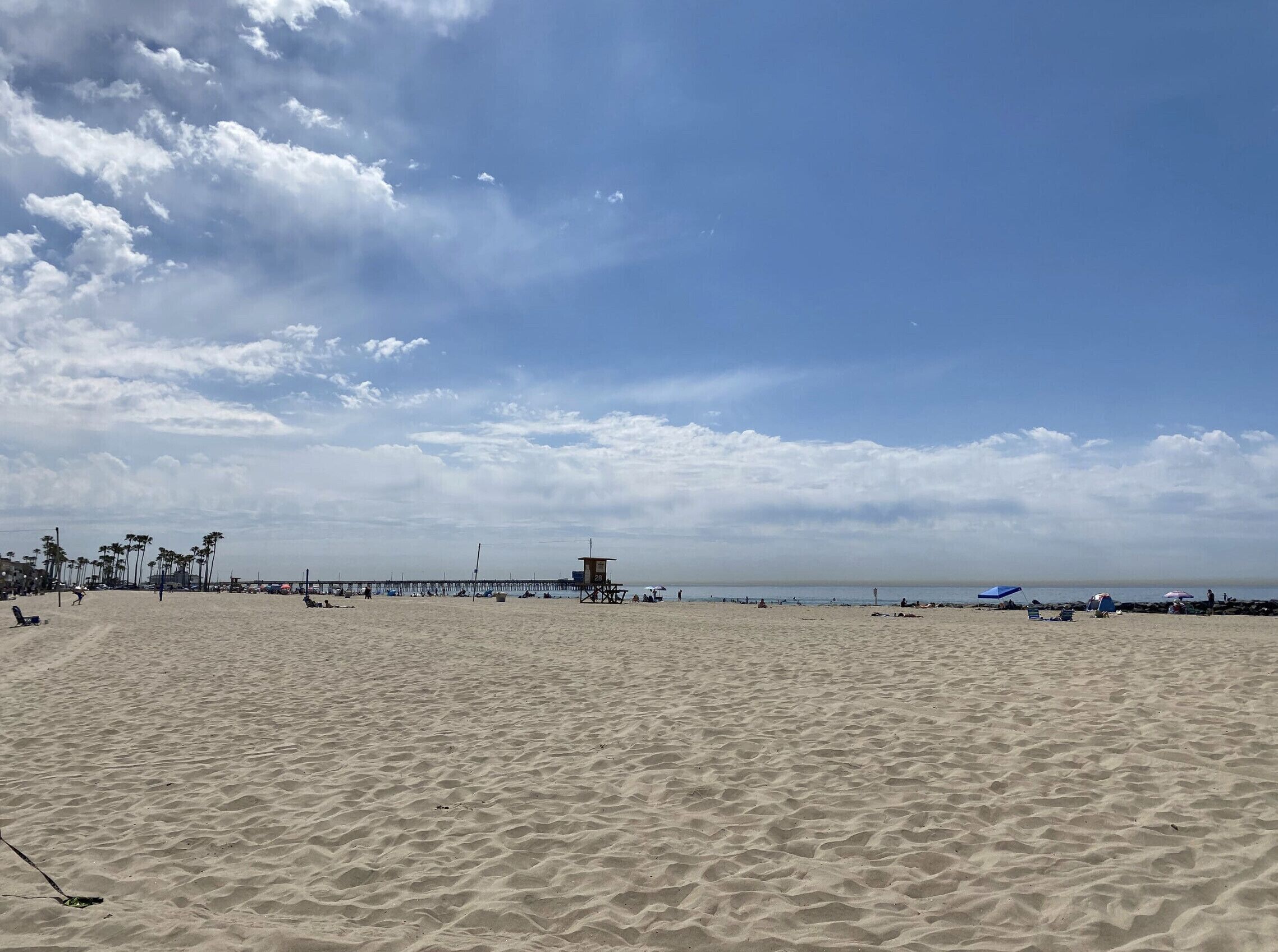 A wide sandy beach with a pier extending into the water, palm trees, and a lifeguard tower.