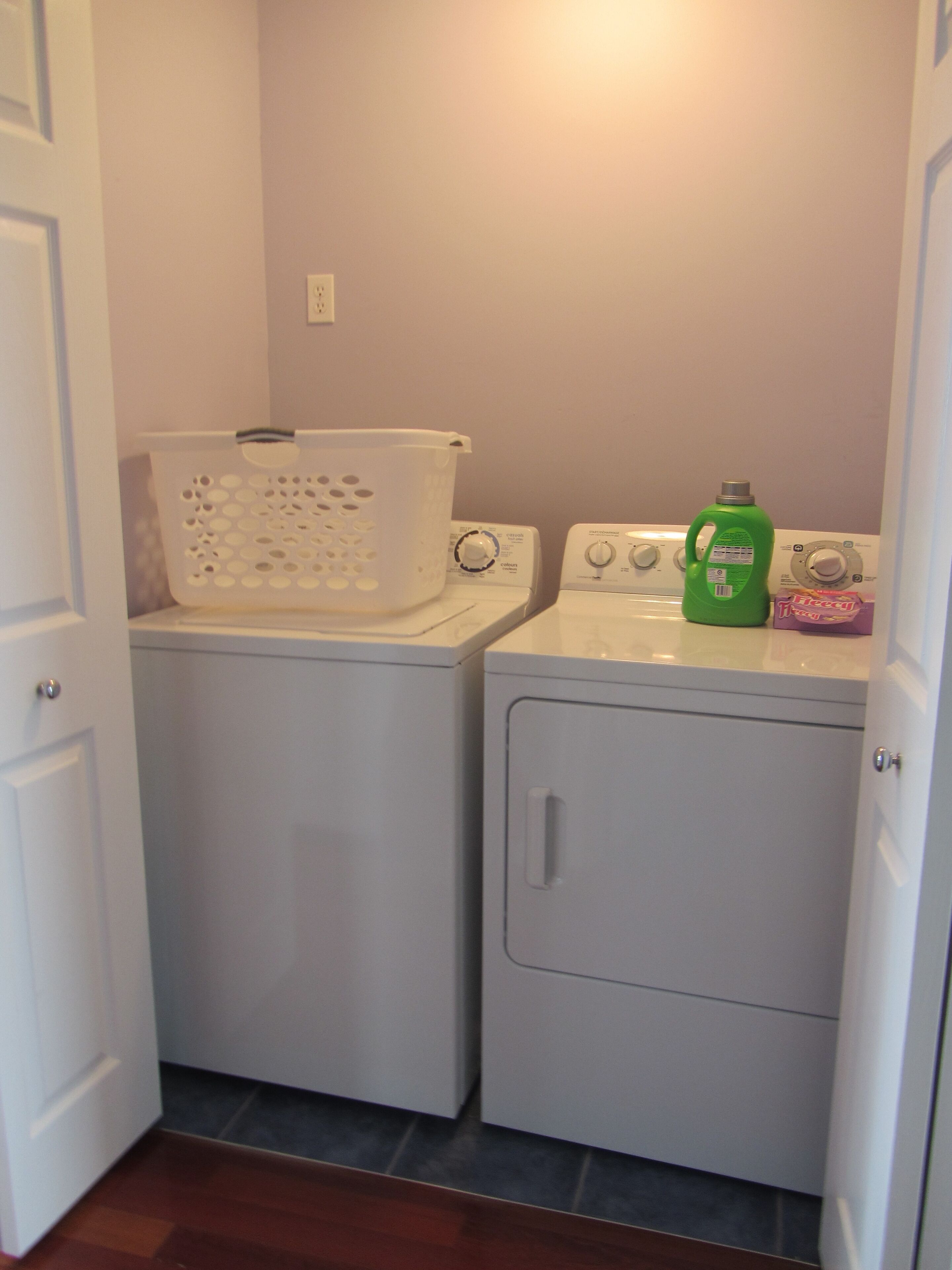 A laundry area with two washing machines, a white laundry basket, and a green detergent bottle.