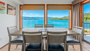 Zona pranzo con tavolo e sedie, ampie finestre con vista su un lago e montagne, e un balcone con vista.