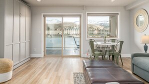 A spacious room with a large sliding glass door leading to a balcony with a view of mountains and a resort. There is a dining area with a table and chairs, and a cushioned bench. - Grandview 627 Waterfront condo on Lake Chelan (Chelan)