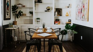 A dining area with a wooden table set for two, surrounded by black chairs. There are shelves with various decorative items and plants. - The Phx Bungalow located in north-central phoenix. (Phoenix)