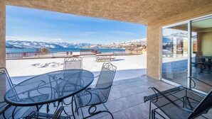 Une terrasse avec une table ronde et des chaises, une porte vitrée donnant sur un espace intérieur, et une vue sur une montagne enneigée et une plage.