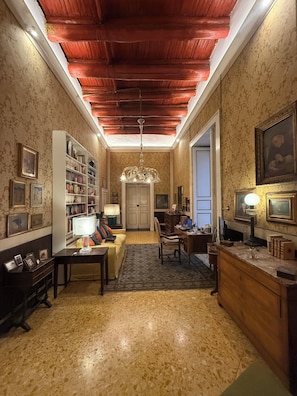 A room with a wooden ceiling, a chandelier, a bookshelf, a desk, a chair, and a door.
