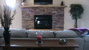 A living room with a stone fireplace, a flat-screen TV mounted above it, a sofa, a coffee table with a vase and a candle, and a potted plant. - Luxury and location, King & Queen pillow, fireplace, whirlpool tub (Frisco)