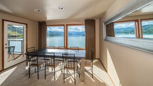 Un coin repas avec une table et des chaises, de grandes fenêtres offrant une vue sur un lac et des montagnes, et une porte donnant sur un balcon.