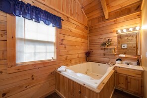 A wooden cabin bathroom with a large bathtub, a vanity with a mirror, and a window with blinds.