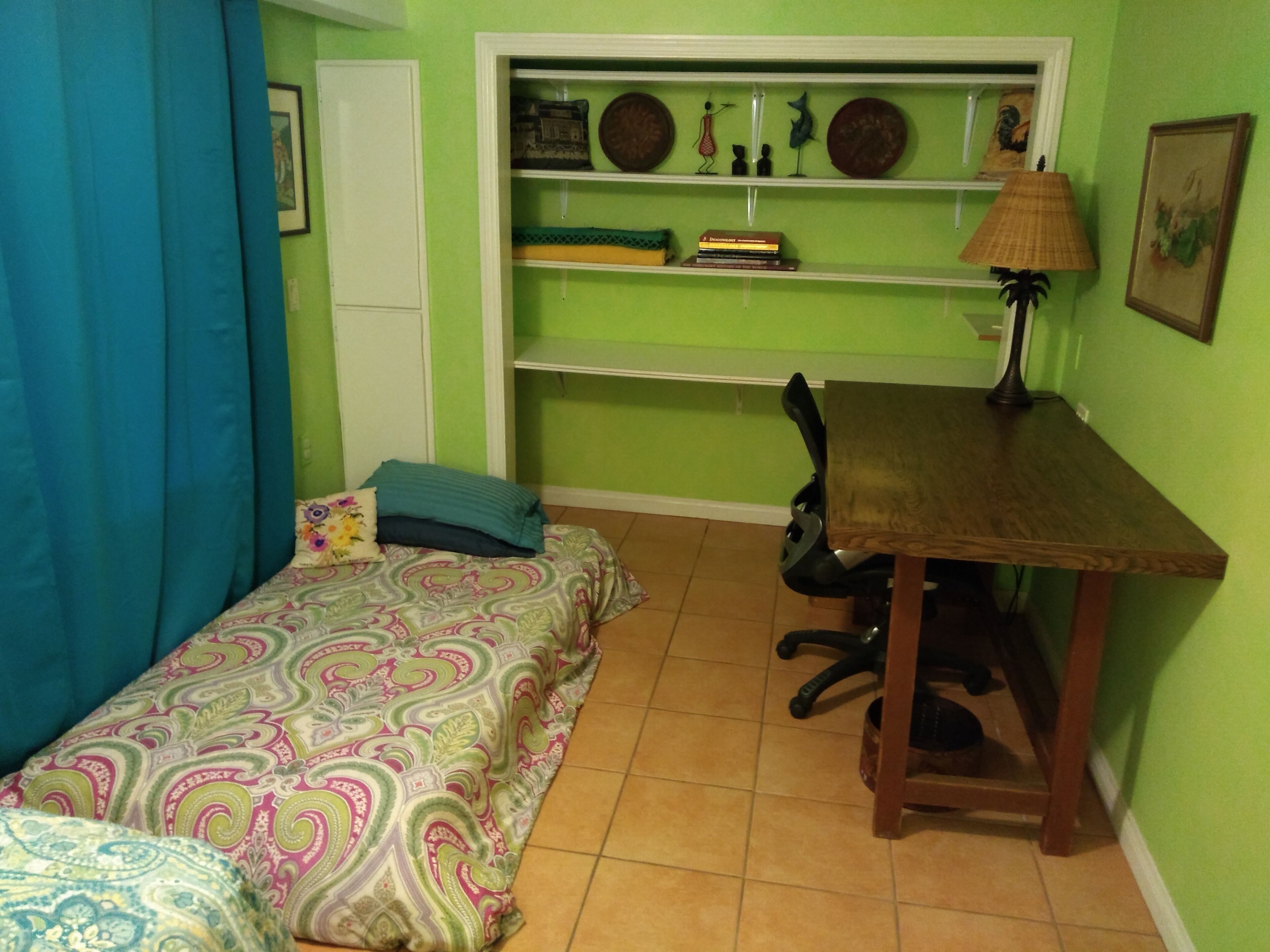 A small bedroom with a bed, a desk, and a chair. There is a shelf above the desk with decorative items.