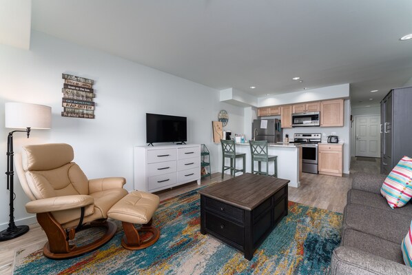 A modern living room with a beige leather chair, a black ottoman, and a wooden coffee table. A flat-screen TV is mounted on the wall, and a colorful rug is on the floor.