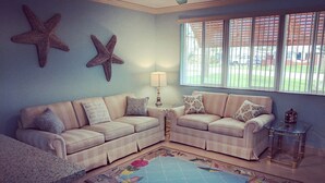 A living room with two beige sofas, starfish wall decor, a patterned rug, and a window with blinds. - Super Cute Beach Home *Recommended** Casa Azalea (Ormond Beach)