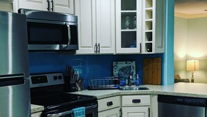 A kitchen with white cabinets, a stainless steel refrigerator, a microwave, an oven, and a dishwasher. There is a blue backsplash, a dish rack, and a placemat with text on the floor. - Super Cute Beach Home *Recommended** Casa Azalea (Ormond Beach)