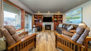 A spacious living room with a fireplace, wooden furniture, and a large window offering a view of the outdoors. - GrandView #102, 2-Bedroom Waterfront Condo (Chelan)