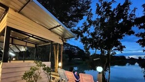 Une terrasse en bois, agrémentée de plantes en pot et de chaises en plastique, donnant sur une étendue d’eau calme au crépuscule.