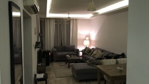 Un salon moderne avec un canapé, un pouf et une table à manger. On y trouve une lampe, un luminaire au plafond et un appareil de climatisation.