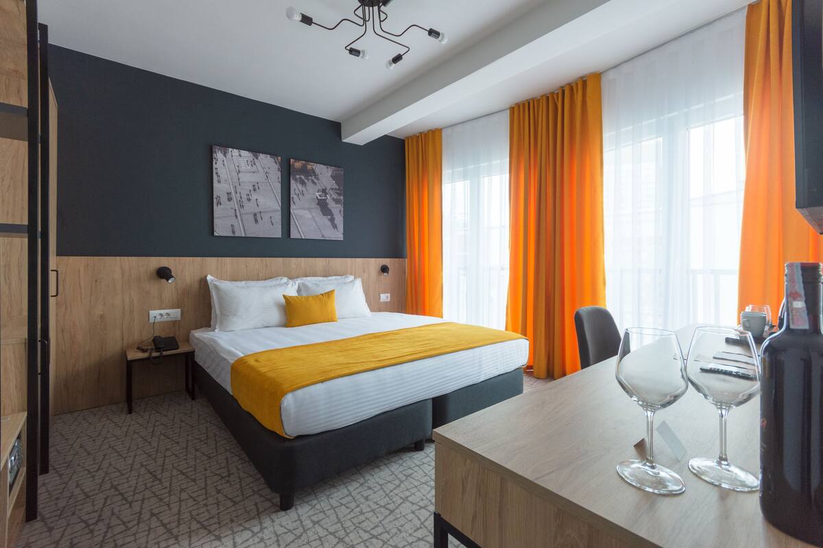 A hotel room with a bed, a desk with two wine glasses, a bottle, and a chair.